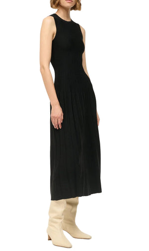 A woman wears the Elyse Dress by Staud—a sleeveless black midi with a fitted bodice, pleated skirt, and crew neckline—styled with off-white knee-high boots.