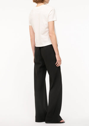 A person stands facing away, wearing a short-sleeve white t-shirt and the Staud Stroll Pant—loose black straight-leg pants with an elastic waistband—against a plain white background.