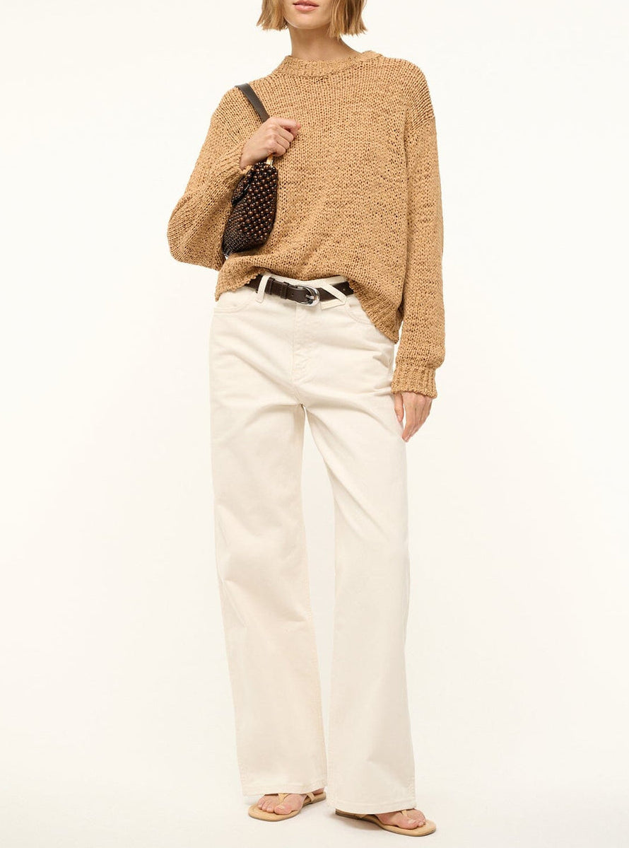 A person stands against a plain light background, wearing a loose tan sweater, white wide-leg pants, beige sandals, and carrying the Staud Timmy Convertible Shoulder Bag—a small black bag with a detachable handle.