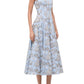 A woman wears the Staud Wells Dress Sky Toile, a light blue cotton poplin midi with a fitted waist and square neckline, paired with black sandals, standing against a white background.