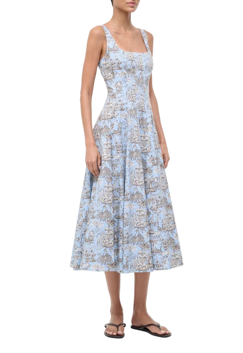 A woman wears the Staud Wells Dress Sky Toile, a light blue cotton poplin midi with a fitted waist and square neckline, paired with black sandals, standing against a white background.