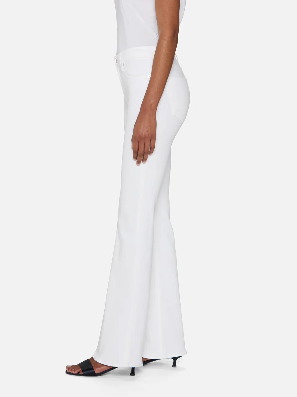 A person stands in profile against a plain background, wearing The Icon high-waisted flare jeans by Frame in white for a '70s-inspired silhouette, styled with a fitted white top and black open-toe heeled sandals.