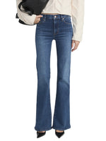 A person wearing Frame's The Icon blue ultra-stretch denim bootcut jeans, a cream textured sweater, black pointed-toe shoes, and holding a black bag.