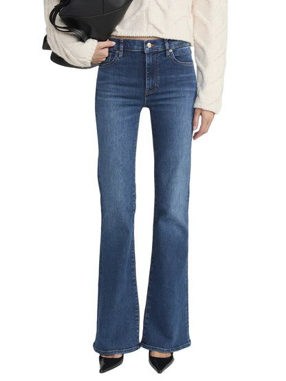 A person wearing Frame's The Icon blue ultra-stretch denim bootcut jeans, a cream textured sweater, black pointed-toe shoes, and holding a black bag.