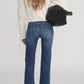 A person stands with their back to the camera, wearing Frame's The Icon cream knit sweater, blue flare jeans made from ultra-stretch denim, and holding a black bag under one arm.