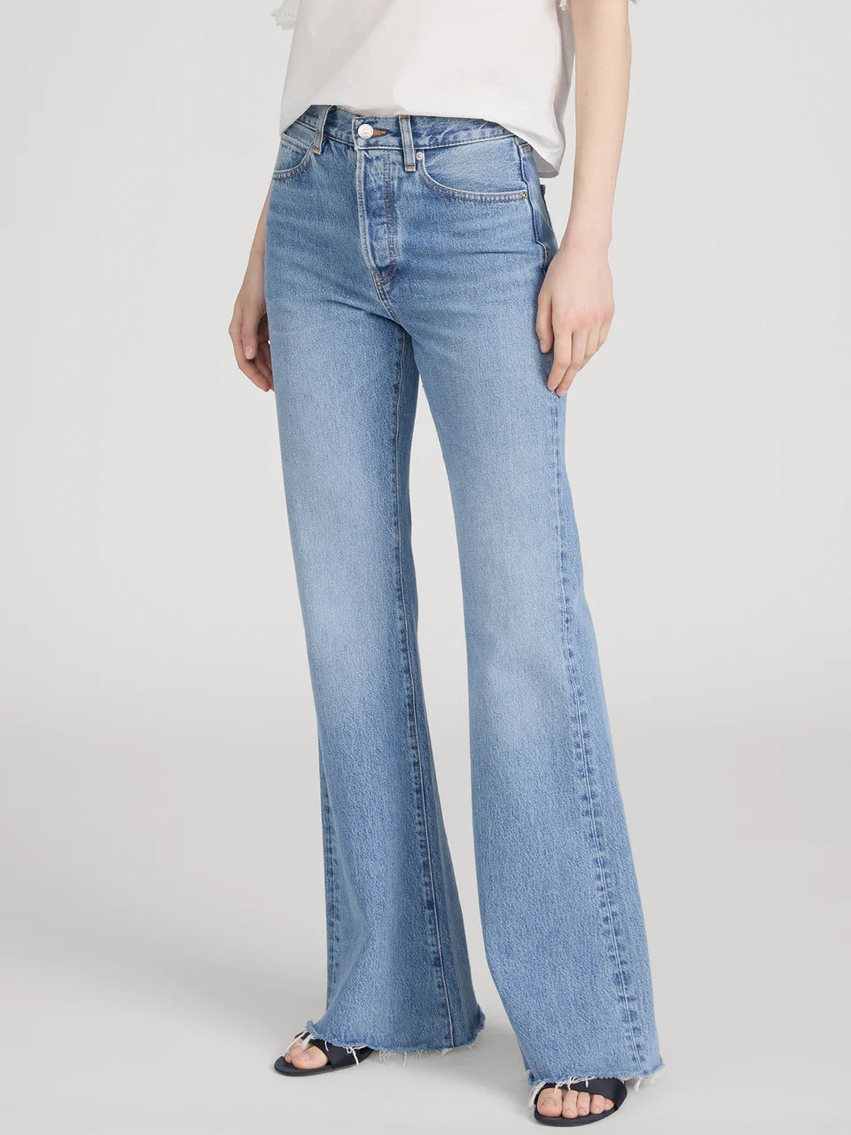A person wearing Frame's The Lax rigid denim jeans—light blue, high-waisted, wide-leg flare style with frayed hems—paired with a white short-sleeve top and black sandals, stands against a plain background for a vintage vibe.