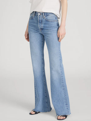 A person wearing Frame's The Lax rigid denim jeans—light blue, high-waisted, wide-leg flare style with frayed hems—paired with a white short-sleeve top and black sandals, stands against a plain background for a vintage vibe.