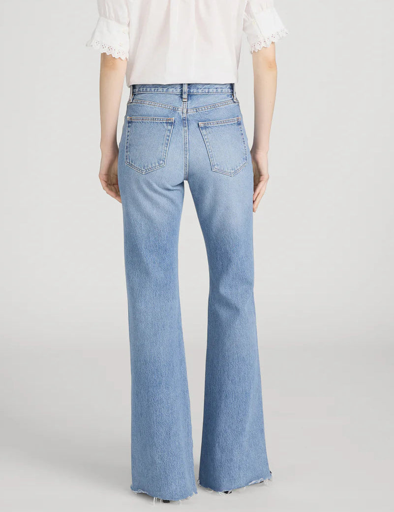 Model wears The Lax by Frame: a white short-sleeve blouse paired with high-waisted, light blue wide-leg flare jeans in classic rigid denim, shown from the back against a plain background for a vintage vibe.