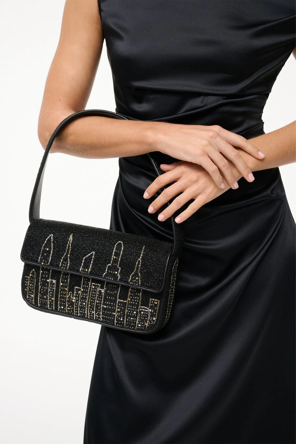 A woman in a black satin dress holds the Staud Tommy Beaded Bag, a vegan leather handbag adorned with a gold city skyline design.