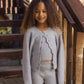 A young girl with curly hair stands by wooden stairs, wearing the Rylee & Cru Girls’ Fuzzy Knit Lounge set in light grey, featuring a matching ribbed cardigan, top, and wide leg pants.