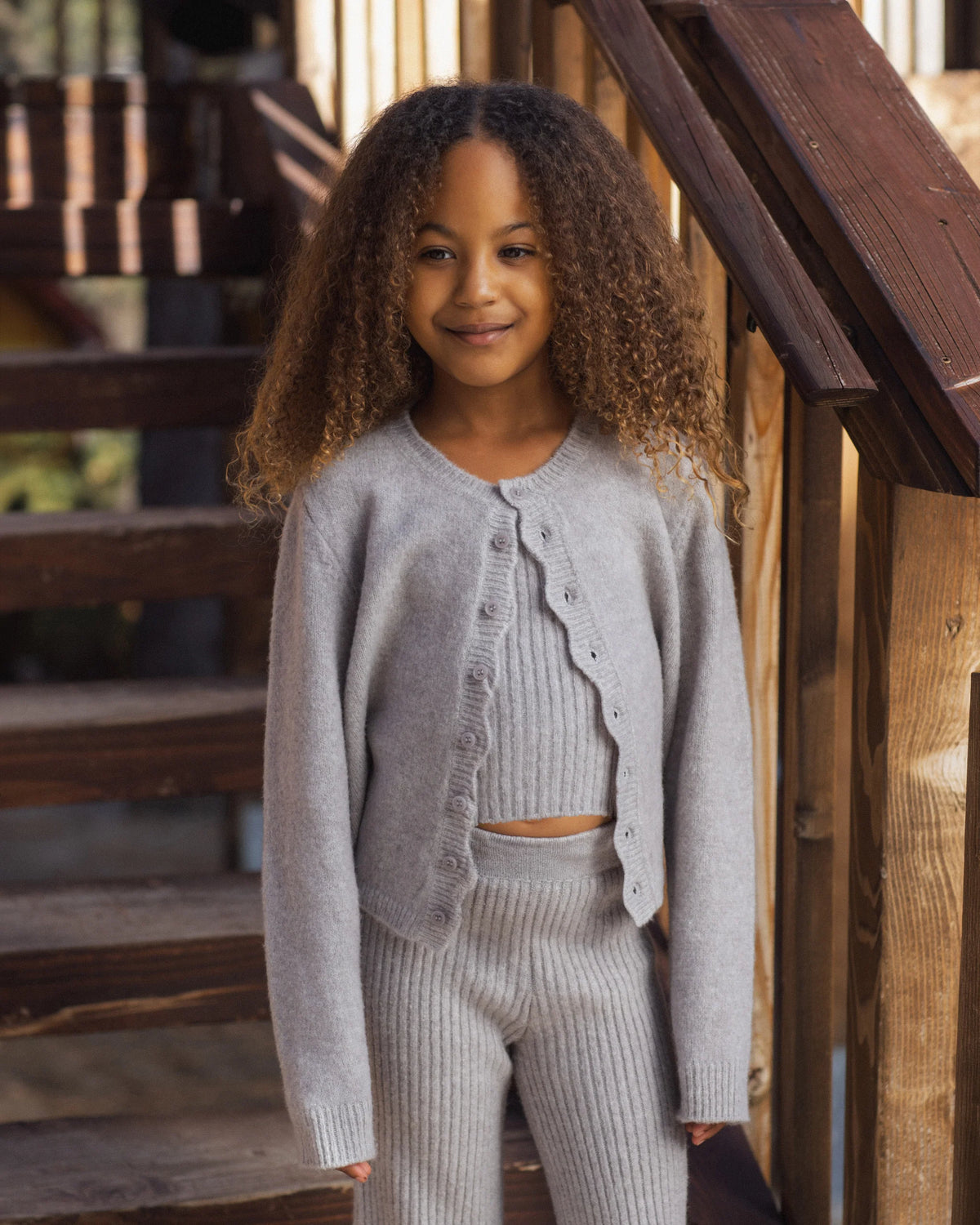 A young girl with curly hair stands by wooden stairs, wearing the Rylee & Cru Girls’ Fuzzy Knit Lounge set in light grey, featuring a matching ribbed cardigan, top, and wide leg pants.