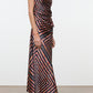 A woman wears the Staud Portia Silk Studio Dress, an asymmetrical silk midi dress with a sleeveless design and diagonal geometric stripes in black, orange, and pink. The look is paired with black low-heeled shoes. The dress features an adjustable tie detail at the waist and the woman stands on a plain floor against a white wall.