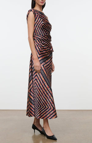 A woman wears the Staud Portia Silk Studio Dress, an asymmetrical silk midi dress with a sleeveless design and diagonal geometric stripes in black, orange, and pink. The look is paired with black low-heeled shoes. The dress features an adjustable tie detail at the waist and the woman stands on a plain floor against a white wall.