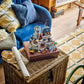 The LEGO® Creator™ 3-in-1 Medieval Horse Knight Castle by Legos - Toyhouse sits atop a closed book on a wicker basket, with a sofa and patterned rug in the living room background.