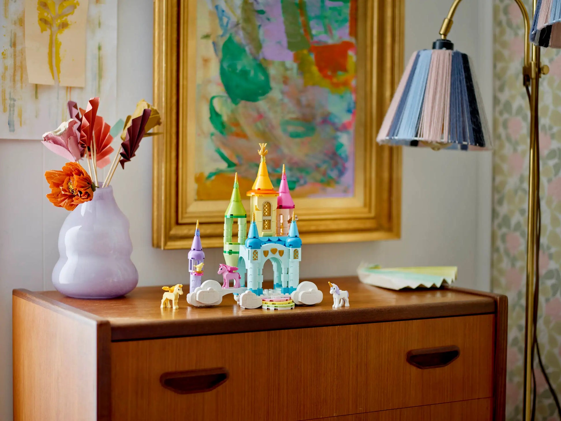 The LEGO® Creator™ 3-in-1 Unicorn Castle by Legos - Toyhouse, featuring animal figures and a unicorn toy, sits on a wooden dresser beside a pastel flower vase, underneath a framed abstract painting and a lamp.