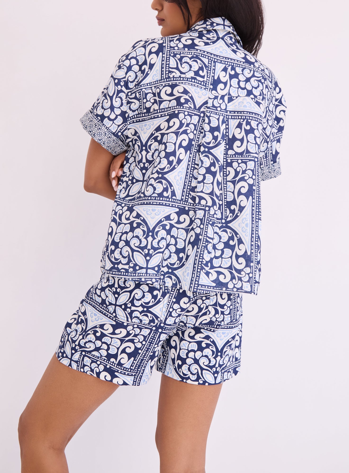 A person wearing the Cara Cara Calvia Top, a blue and white patterned short-sleeve button-down shirt, paired with matching shorts, stands against a plain light background.