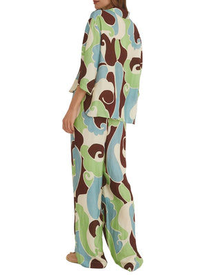 A person is seen from the back wearing Cara Cara's Girona Pant, a flowy drawstring style with gold cord ends, paired with a loose long-sleeve top. The outfit features an abstract green, blue, brown, and cream pattern.