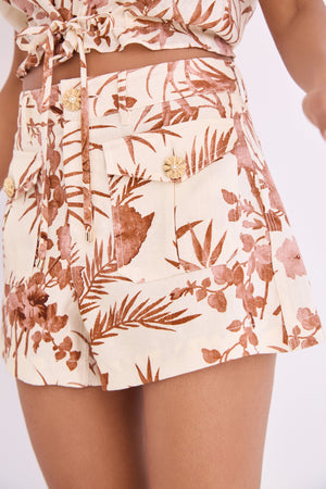 Someone dressed in Cara Cara's Xenia Short, featuring a high-waisted mini cut with a bold brown floral and leaf print and decorative pocket buttons.