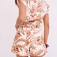 A woman is shown from the back wearing the Cara Cara Xenia Short, a cream short-sleeved romper featuring mini shorts and a bold brown tropical leaf and floral print, set against a plain background.