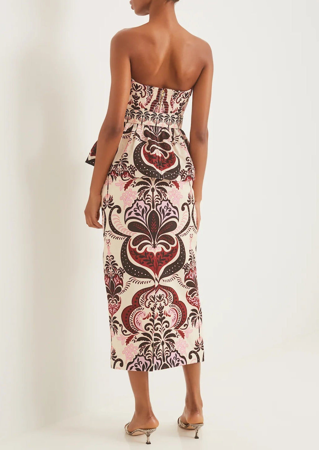 A woman faces away, wearing the Cara Cara Sienna Skirt, an ankle-length piece with a waisted fit and bold red, pink, and brown floral scroll patterns, styled with heeled sandals.