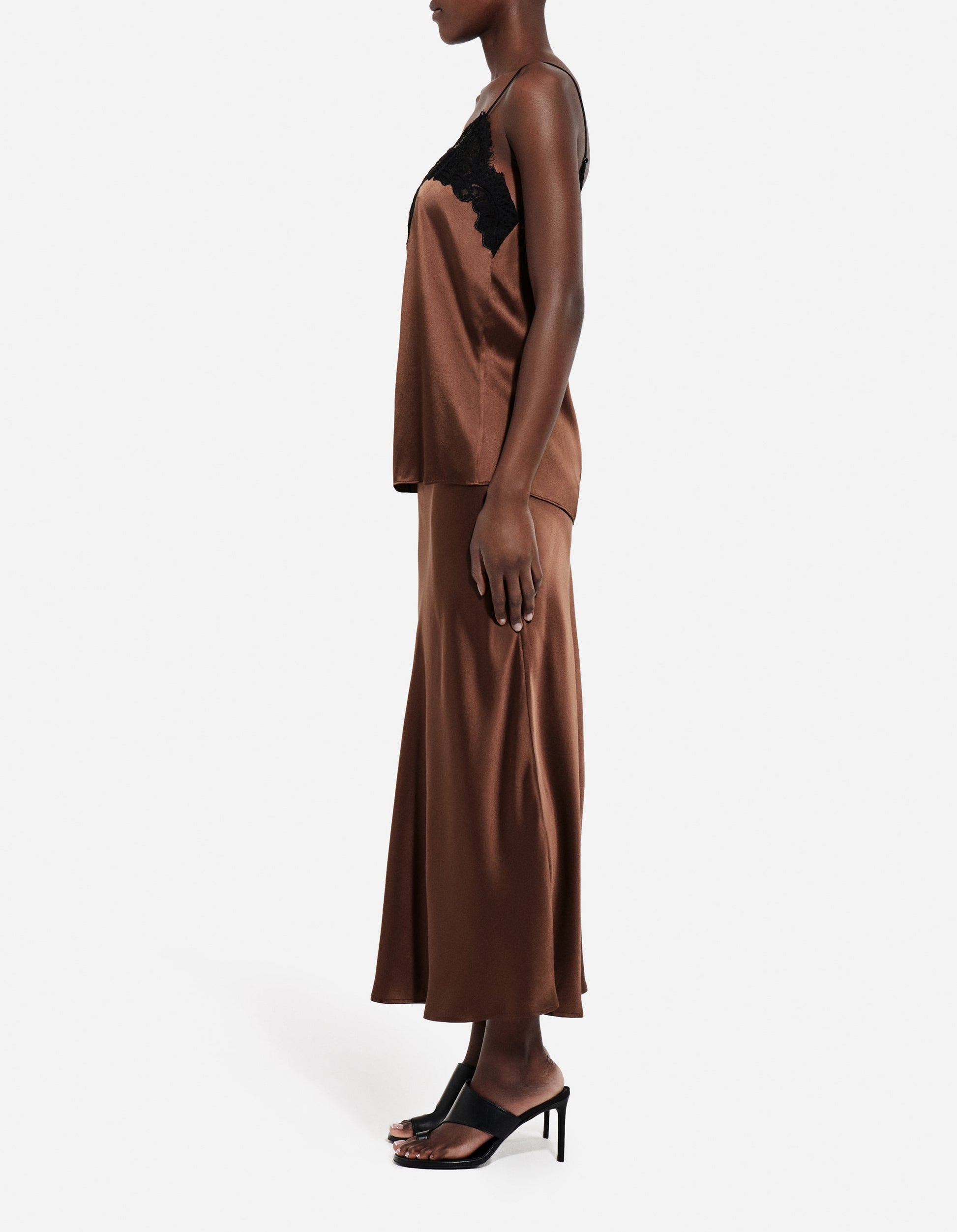 A person stands in profile wearing CO's Slip Silk Skirt, a brown satin bias cut style, paired with a matching camisole featuring black lace trim and black high-heeled sandals.