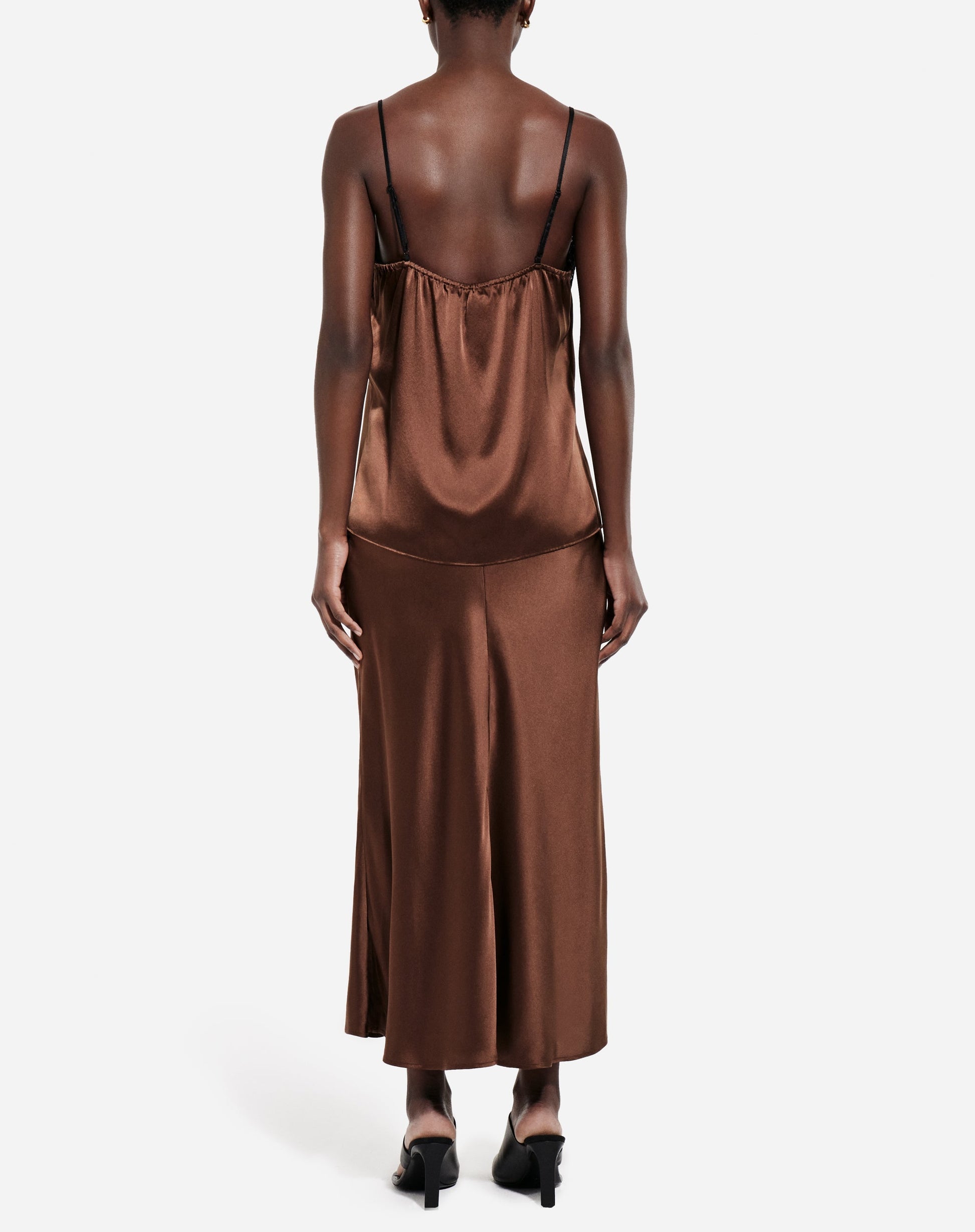 A person is shown from the back against a plain white background, wearing the CO Slip Silk Skirt in brown satin styled as a midi slip dress with thin straps and black heels, evoking the look of a classic bias-cut slip.