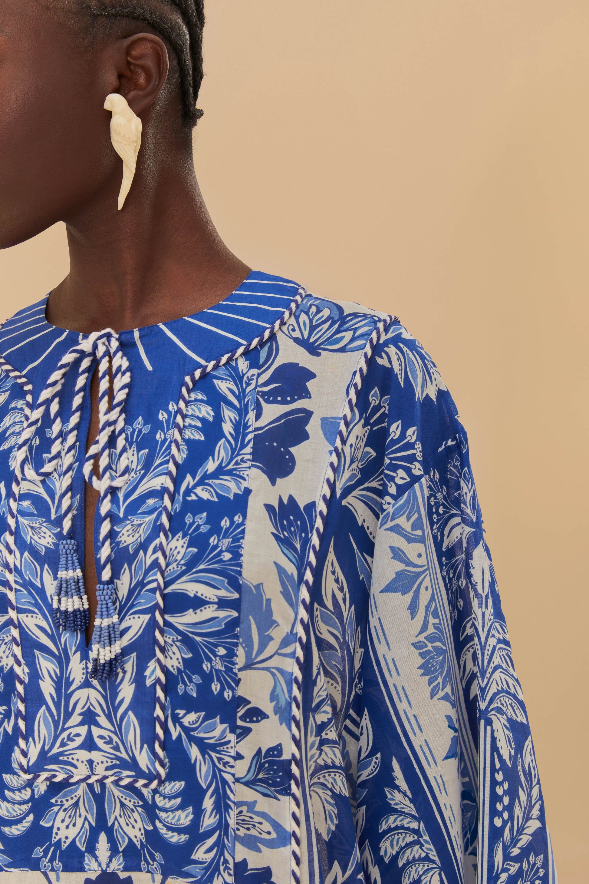 A person wearing the Farm Rio Delicate Tapestry Long Cover Up with botanical patterns and a tie neckline, styled with cream leaf-shaped earrings against an off-white beige background.