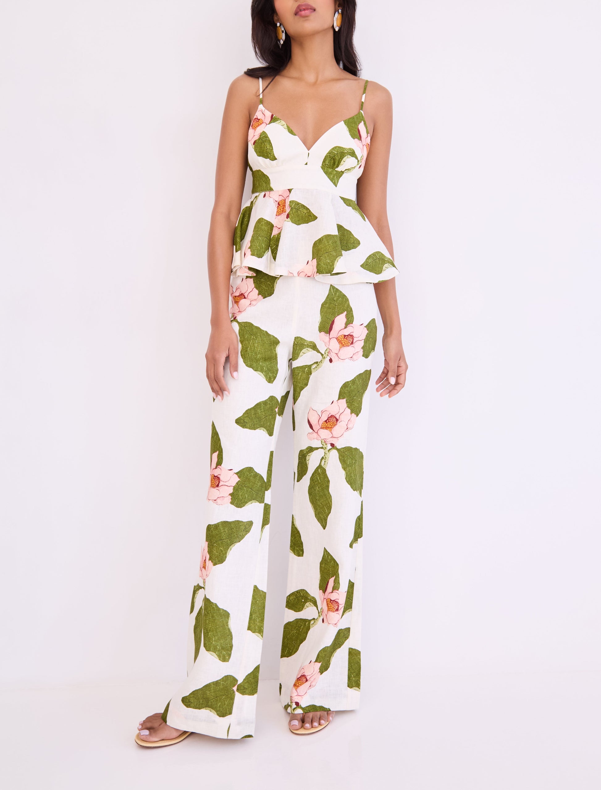 A woman stands against a plain background wearing Cara Cara’s Sunflower Pant, featuring a green and pink floral print, paired with a white sleeveless peplum top for a chic, tailored look.