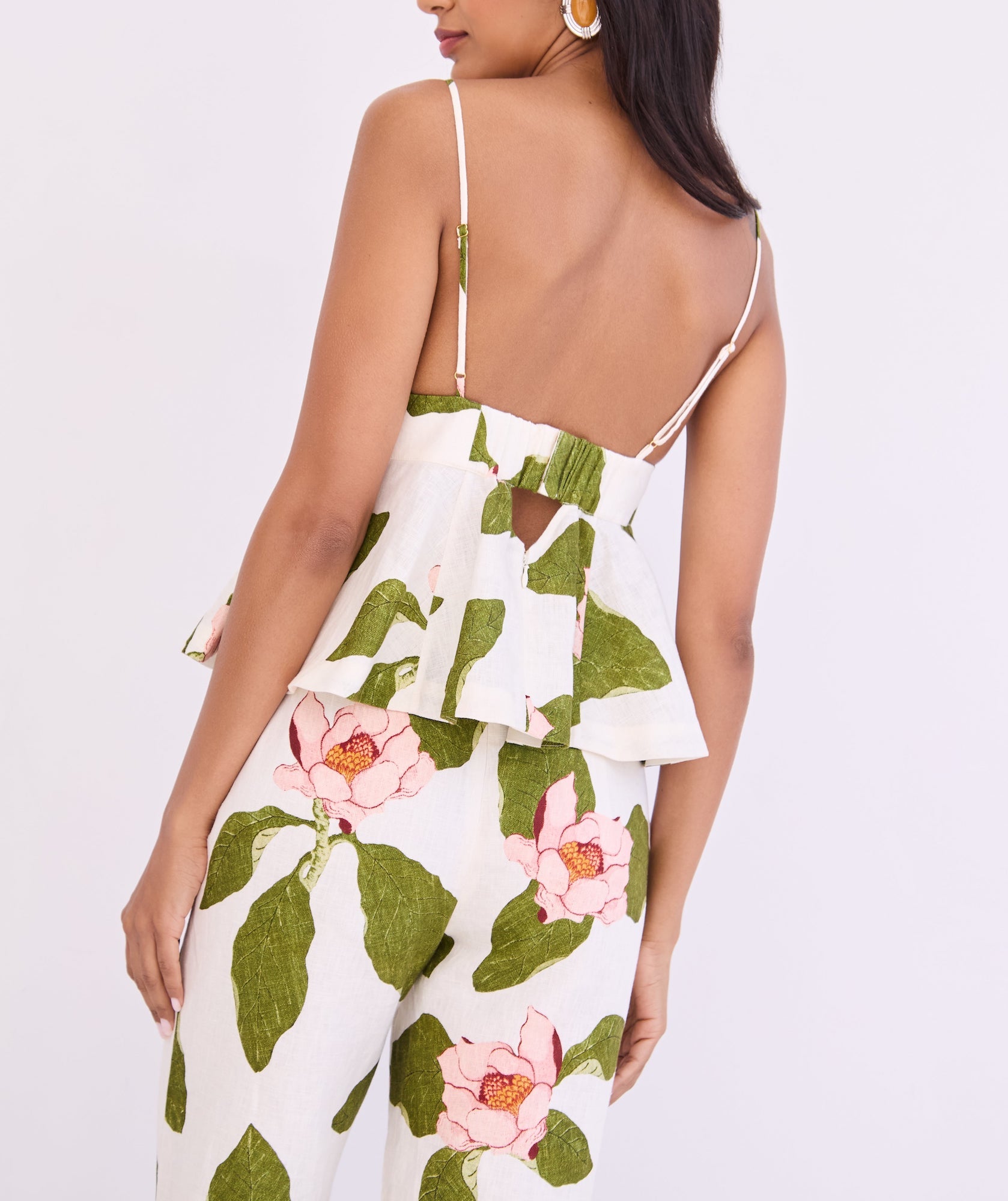 A woman is seen from the back wearing the Cara Cara Avenue Top—a white fitted peplum top with adjustable thin straps, a pink and green floral print, ruffled peplum hem, and a back cutout.