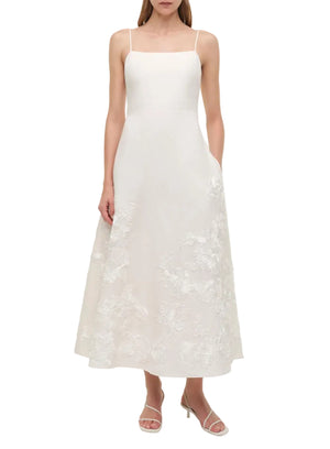 A woman wears the Simkhai Annabella Soutache Dress, a sleeveless white linen-blend midi with thin straps and detailed soutache embroidery, styled with strappy sandals.