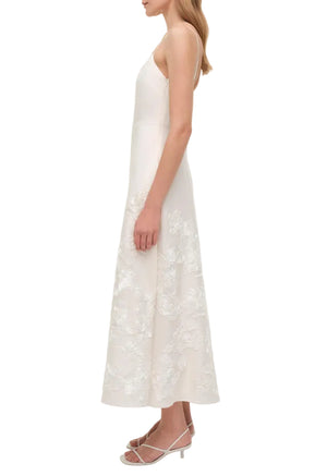 A woman stands in profile wearing the Simkhai Annabella Soutache Dress—a sleeveless white linen-blend midi with floral soutache embroidery—and strappy heeled sandals against a white background.
