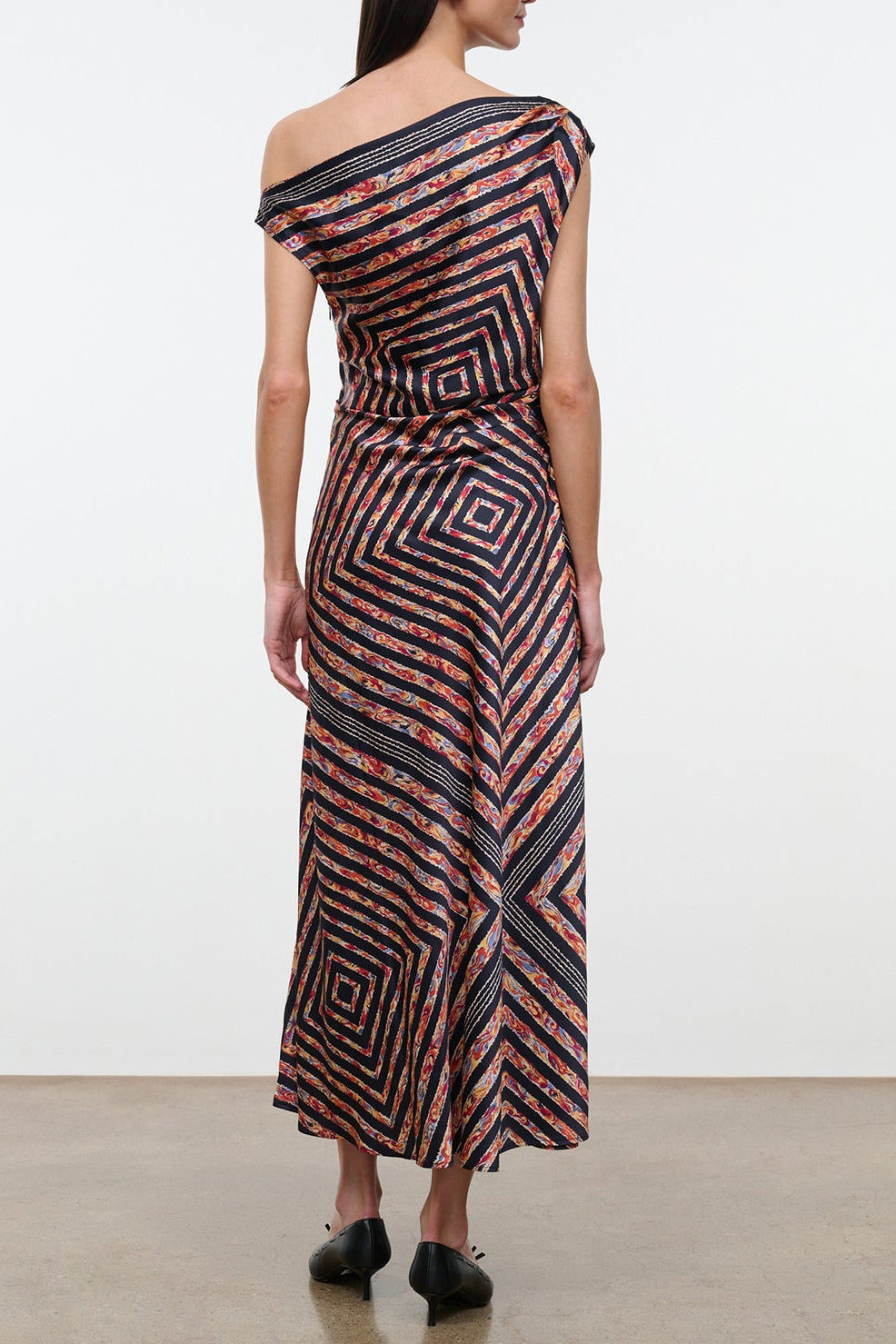 A woman stands with her back to the camera wearing the Staud Portia Silk Studio Dress, an off-shoulder, asymmetrical silk piece adorned with geometric and floral patterns, featuring an adjustable tie detail and paired with black shoes.