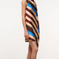 A person stands in profile against a plain background, wearing the Staud Sabina Silk Dress—an easy-fit column dress crafted from printed stretch silk satin and featuring a sleeveless, loose-fitting silhouette with diagonal beige, brown, maroon, and blue stripes. The dress’s asymmetrical design adds an elegant sense of fluidity to the look.