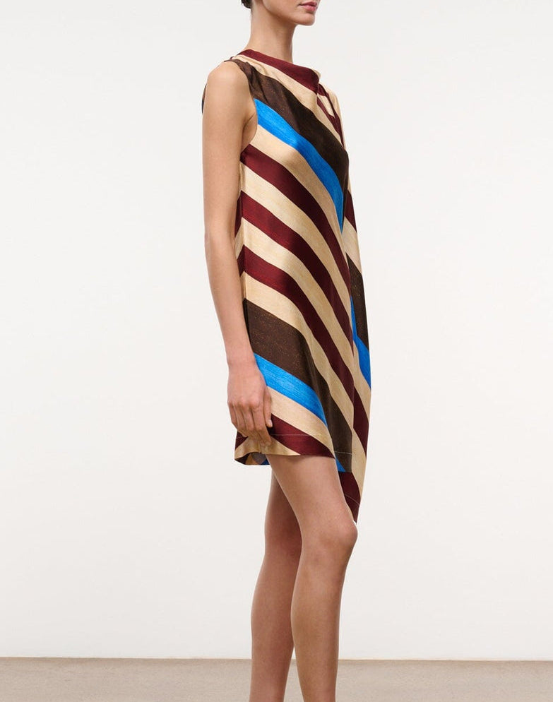 A person stands in profile against a plain background, wearing the Staud Sabina Silk Dress—an easy-fit column dress crafted from printed stretch silk satin and featuring a sleeveless, loose-fitting silhouette with diagonal beige, brown, maroon, and blue stripes. The dress’s asymmetrical design adds an elegant sense of fluidity to the look.