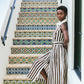A woman in the Cara Cara Quentin Midi Dress, featuring a brown and white collared halter design, stands barefoot beside a staircase adorned with colorful patterned tiles.