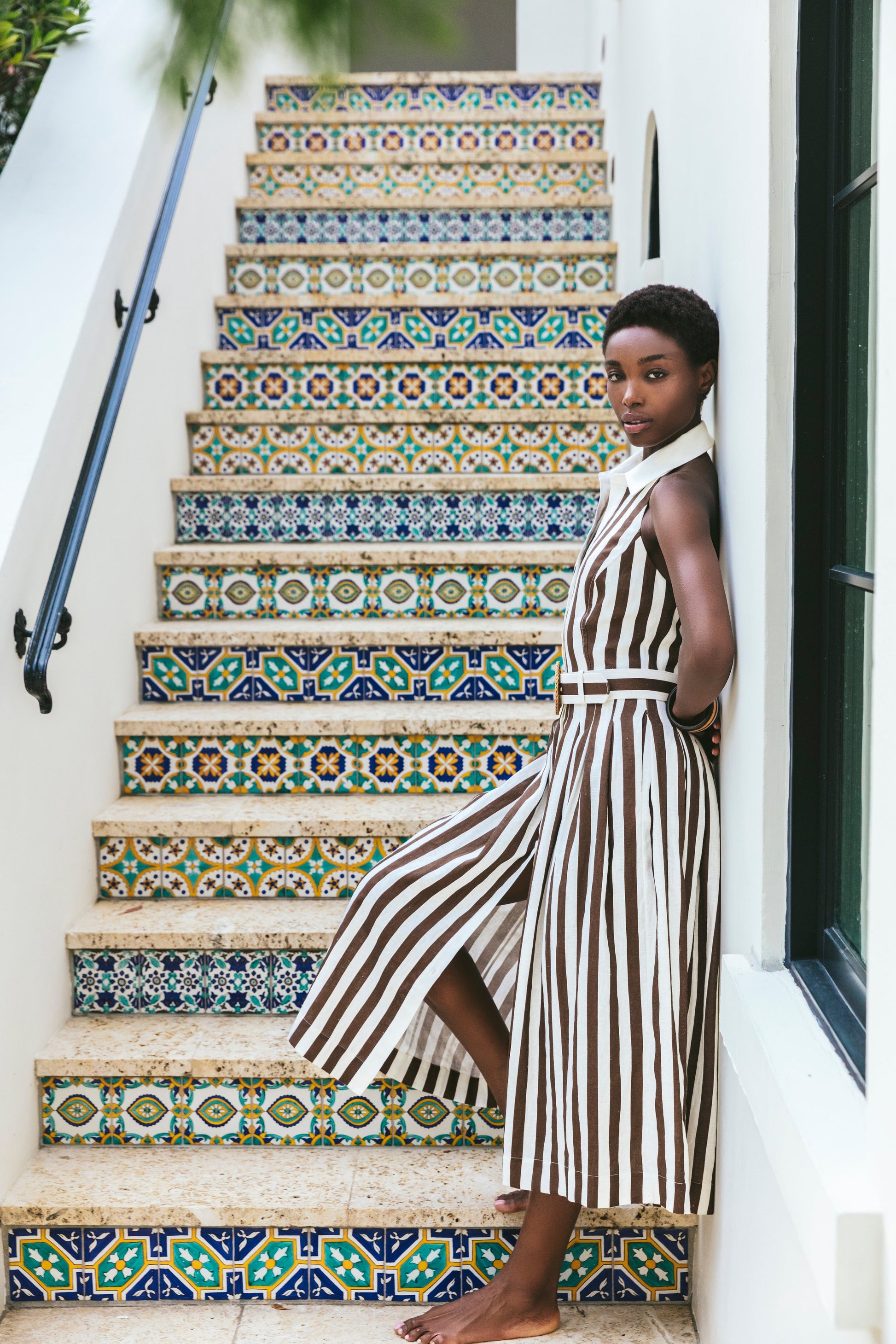 A woman in the Cara Cara Quentin Midi Dress, featuring a brown and white collared halter design, stands barefoot beside a staircase adorned with colorful patterned tiles.