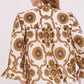 A person wearing the Cara Cara Elowen Top, featuring a cream base with brown and gold floral geometric print, ruffle details on the shoulders, and a picot edge ruffle collar, photographed from the back.