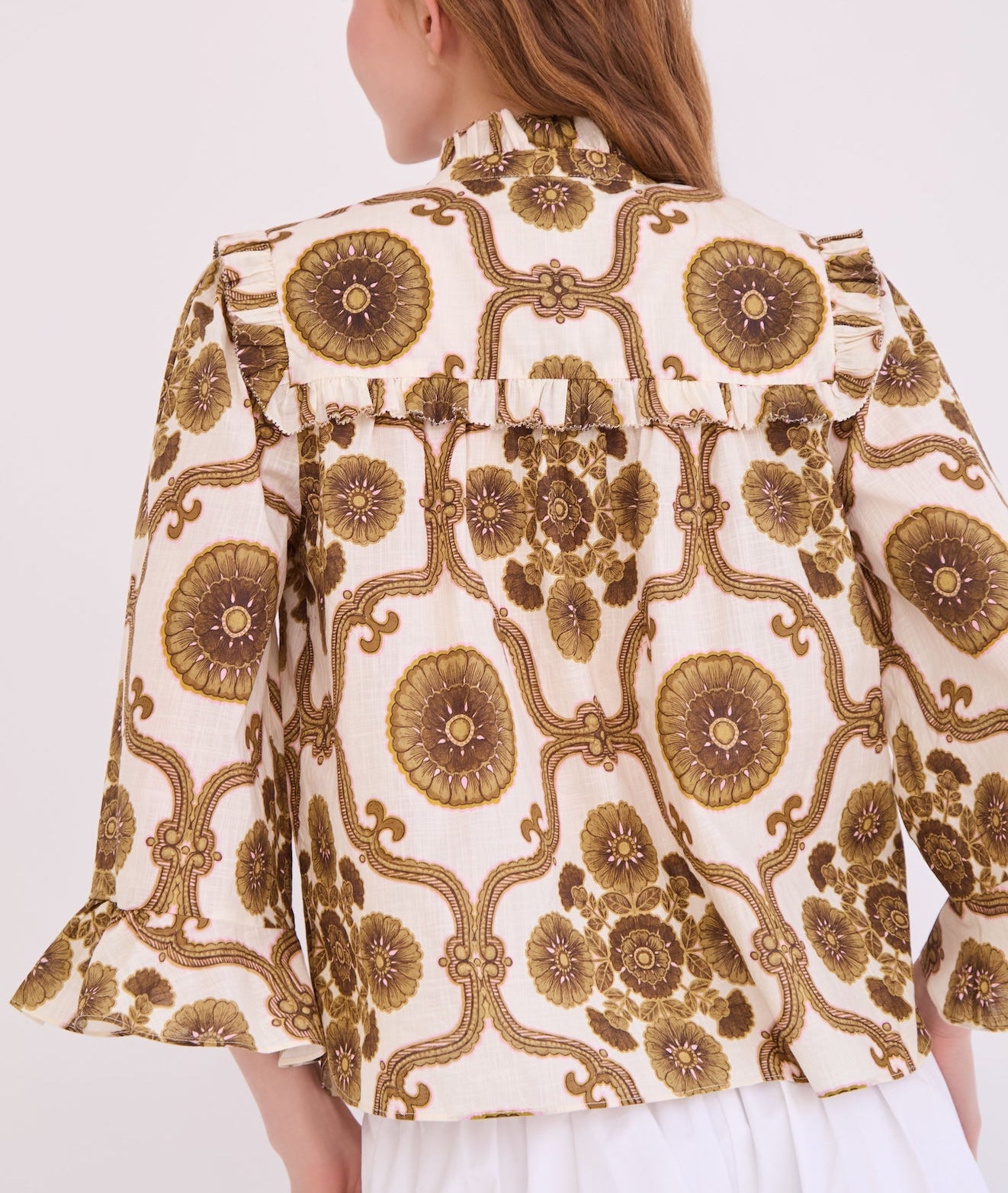 A person wearing the Cara Cara Elowen Top, featuring a cream base with brown and gold floral geometric print, ruffle details on the shoulders, and a picot edge ruffle collar, photographed from the back.