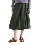 A person wears a gray sweater, TWP's Full Intentions Superfine Cotton Skirt in green with a sculpted silhouette, and brown sandals, standing against a white background.
