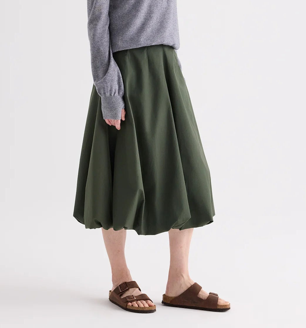 A person wears a grey long-sleeve top, brown sandals, and the TWP Full Intentions Superfine Cotton Skirt in green while standing against a plain background.