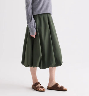 A person wears a grey long-sleeve top, brown sandals, and the TWP Full Intentions Superfine Cotton Skirt in green while standing against a plain background.