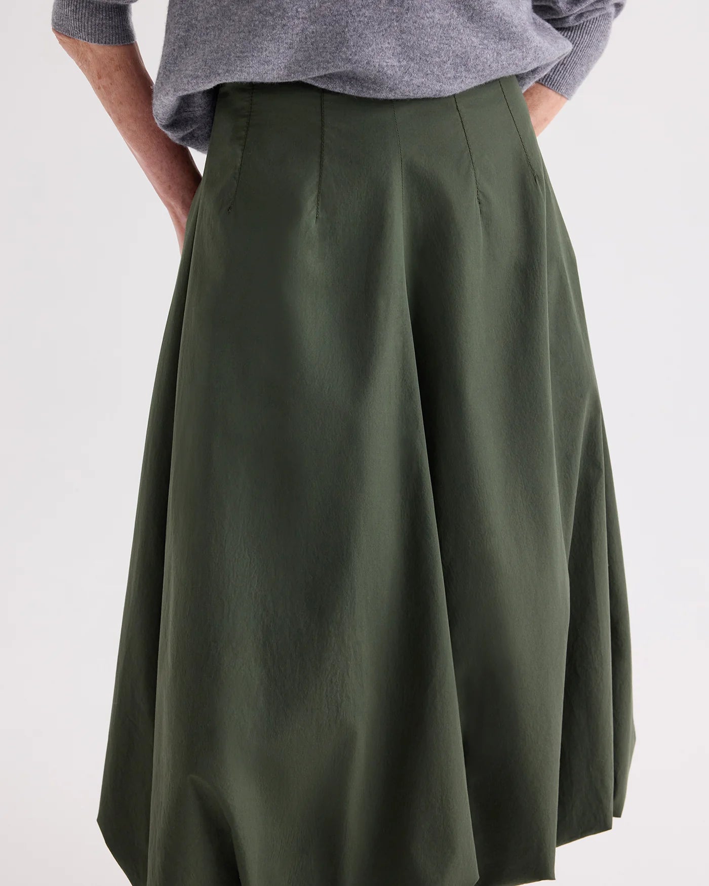 A person wears a gray long-sleeve top tucked into the TWP Full Intentions Superfine Cotton Skirt in olive green, featuring a sculpted silhouette and high waist, with hands resting casually in its pockets.