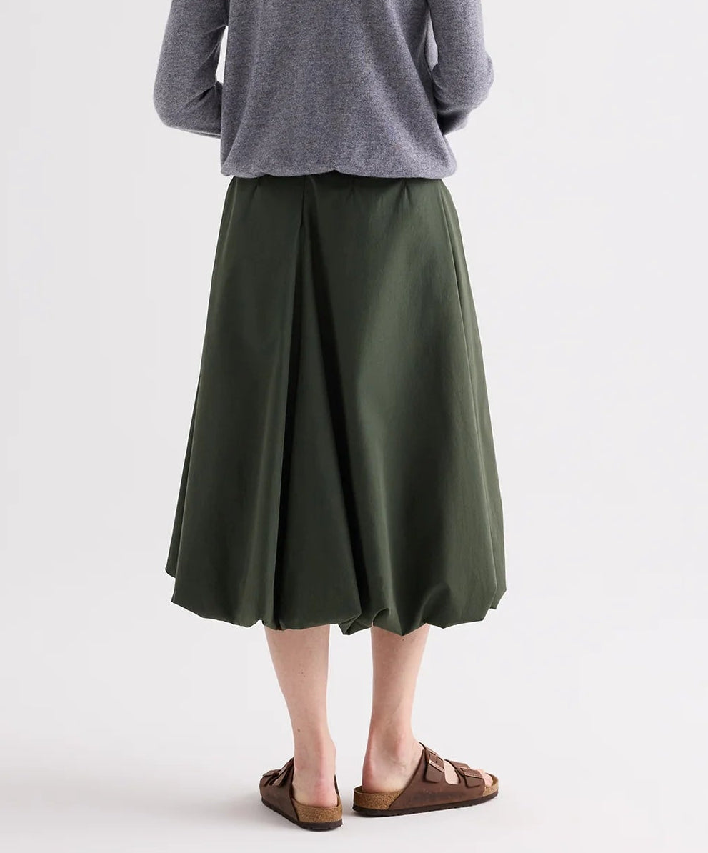 A person stands facing away, wearing a gray long-sleeve top, brown sandals, and the TWP Full Intentions Superfine Cotton Skirt—a voluminous green skirt with a flared hem—against a plain background.