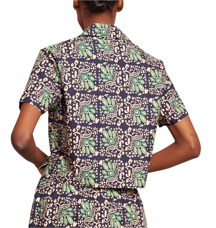 A person stands with hands on hips wearing the Cara Cara Kohut Top, featuring a cropped button-up design with a green and beige paisley Le Stampe print on a dark background, viewed from the back.