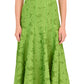 A woman wears the Simkhai Imogen Midi Dress, a strapless green Summer Jacquard style in Kiwi with a floral textured pattern, paired with white open-toe sandals.