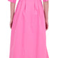 A person is shown from the back wearing Simkhai’s Signature Jazz Dress, a long-sleeved, collared pink cotton poplin midi with an A-line silhouette and fitted waist, paired with white heeled sandals.