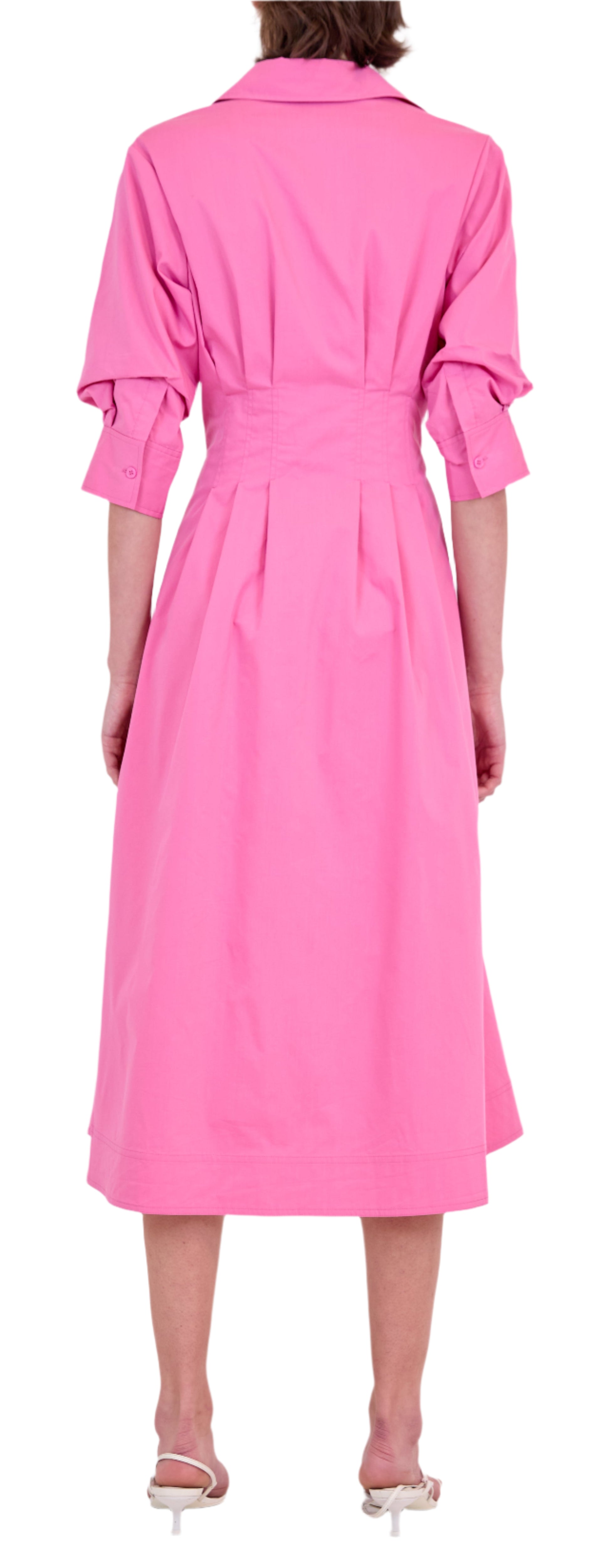 A person is shown from the back wearing Simkhai’s Signature Jazz Dress, a long-sleeved, collared pink cotton poplin midi with an A-line silhouette and fitted waist, paired with white heeled sandals.