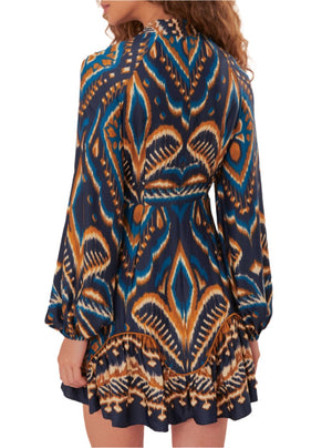 A woman wearing the Farm Rio Pineapple Ikat Blue Mini Dress, a long-sleeve design showcasing a vibrant geometric pattern in shades of orange, blue, and black, complete with a ruffled hem.