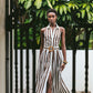 A woman wears the Cara Cara Quentin Midi Dress, featuring brown and white stripes and a belted waist, as she walks on patterned tile with green plants and a white wall in the background.