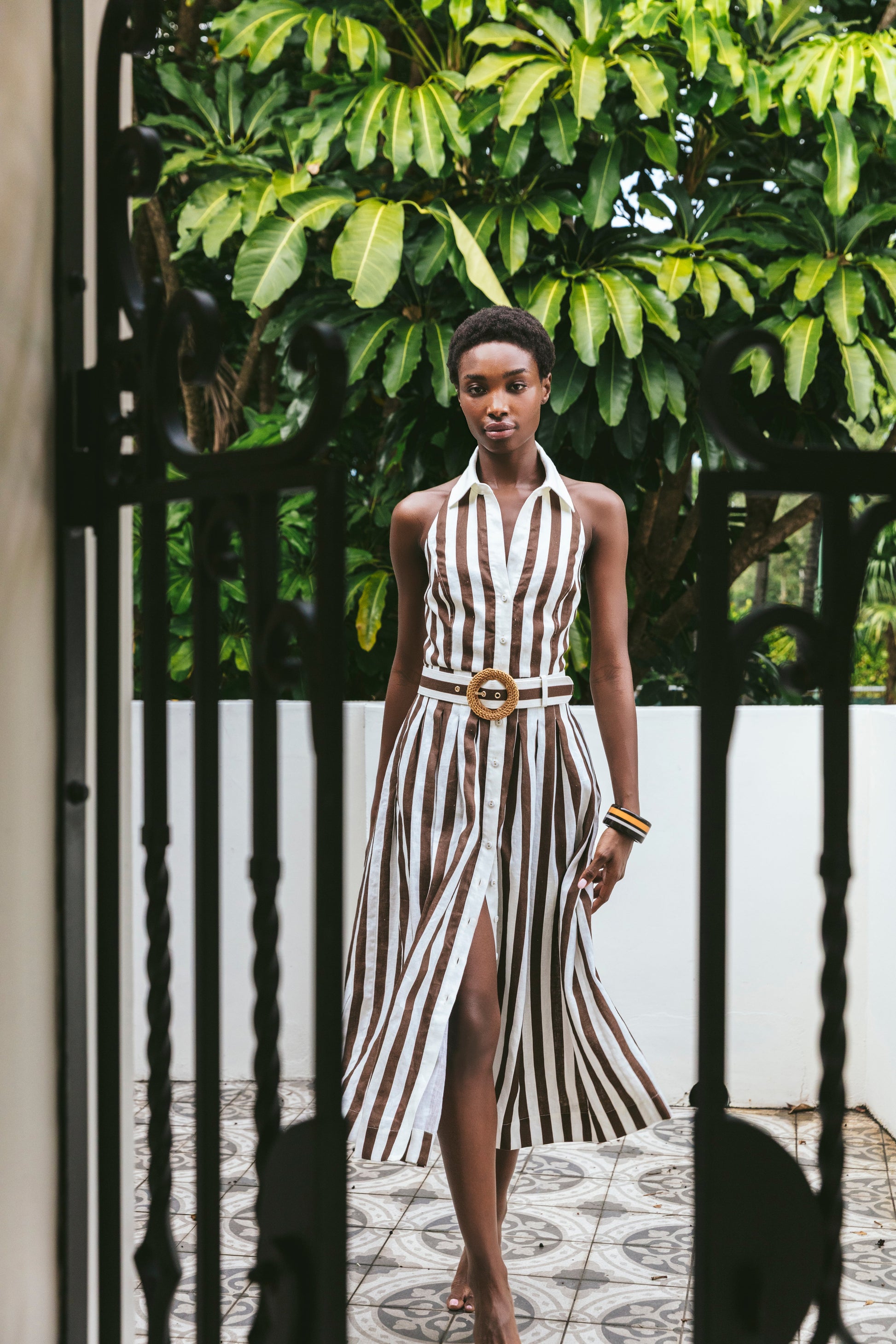 A woman wears the Cara Cara Quentin Midi Dress, featuring brown and white stripes and a belted waist, as she walks on patterned tile with green plants and a white wall in the background.